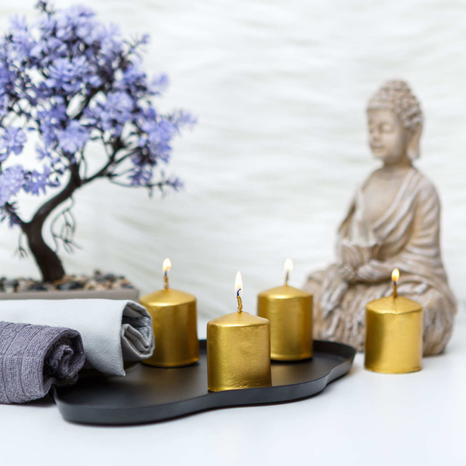 Arany gyertyák fekete tálcán feltekert törölközőkkel és Buddha szoborral a háttérben.