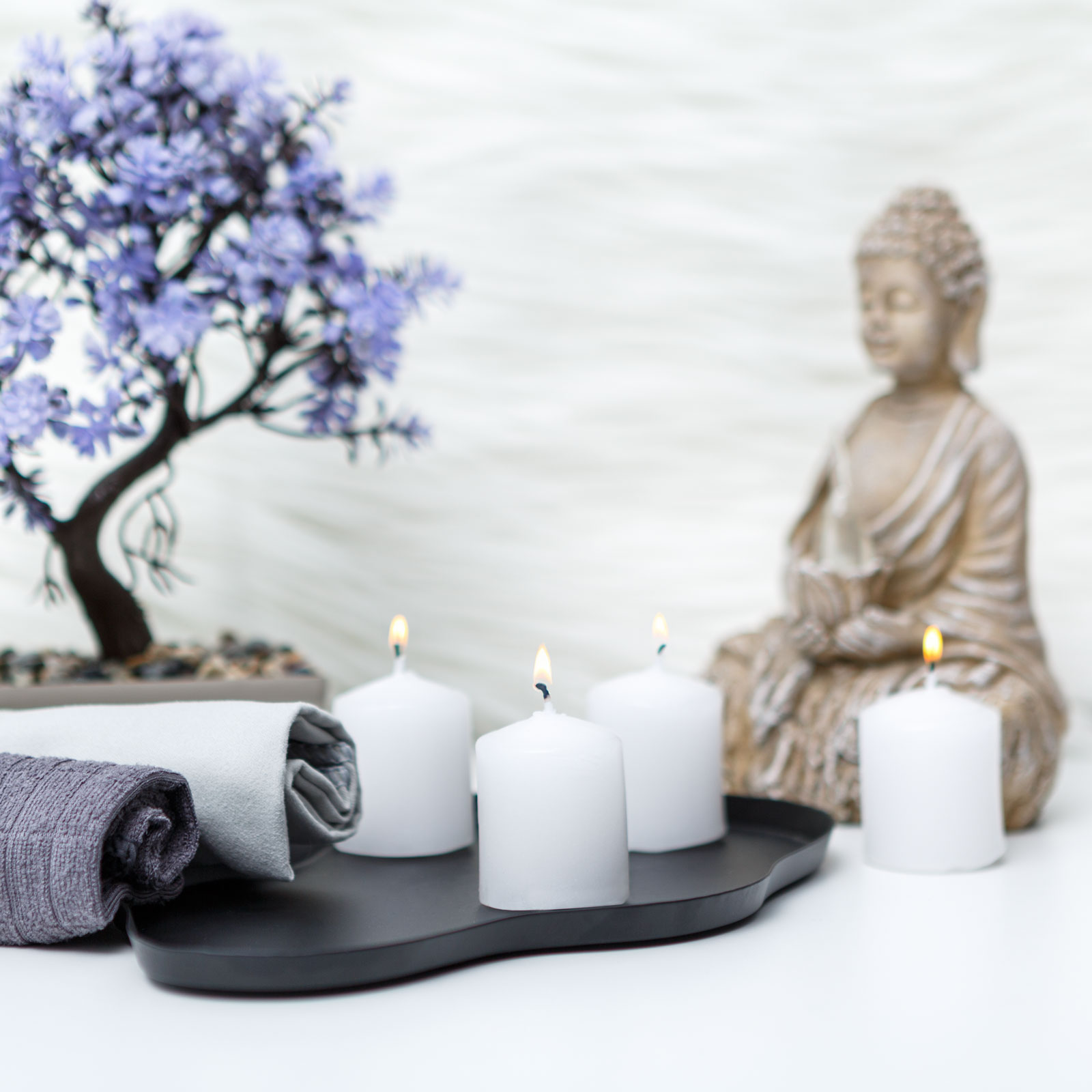 Spa jelenet gyertyákkal, törölközőkkel, bonsai fával és Buddha szoborral. Fehér gyertyák égnek egy fekete tálcán.