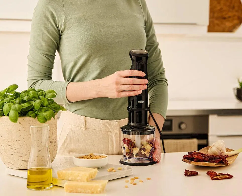 Osoba używająca czarnego blendera. Na stole znajdują się składniki takie jak ser, bazylia, olej i pomidory.