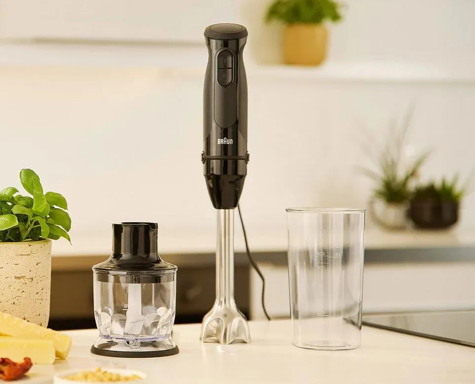 Czarny blender ręczny Braun z akcesoriami na białej powierzchni. Rośliny i jedzenie w tle.