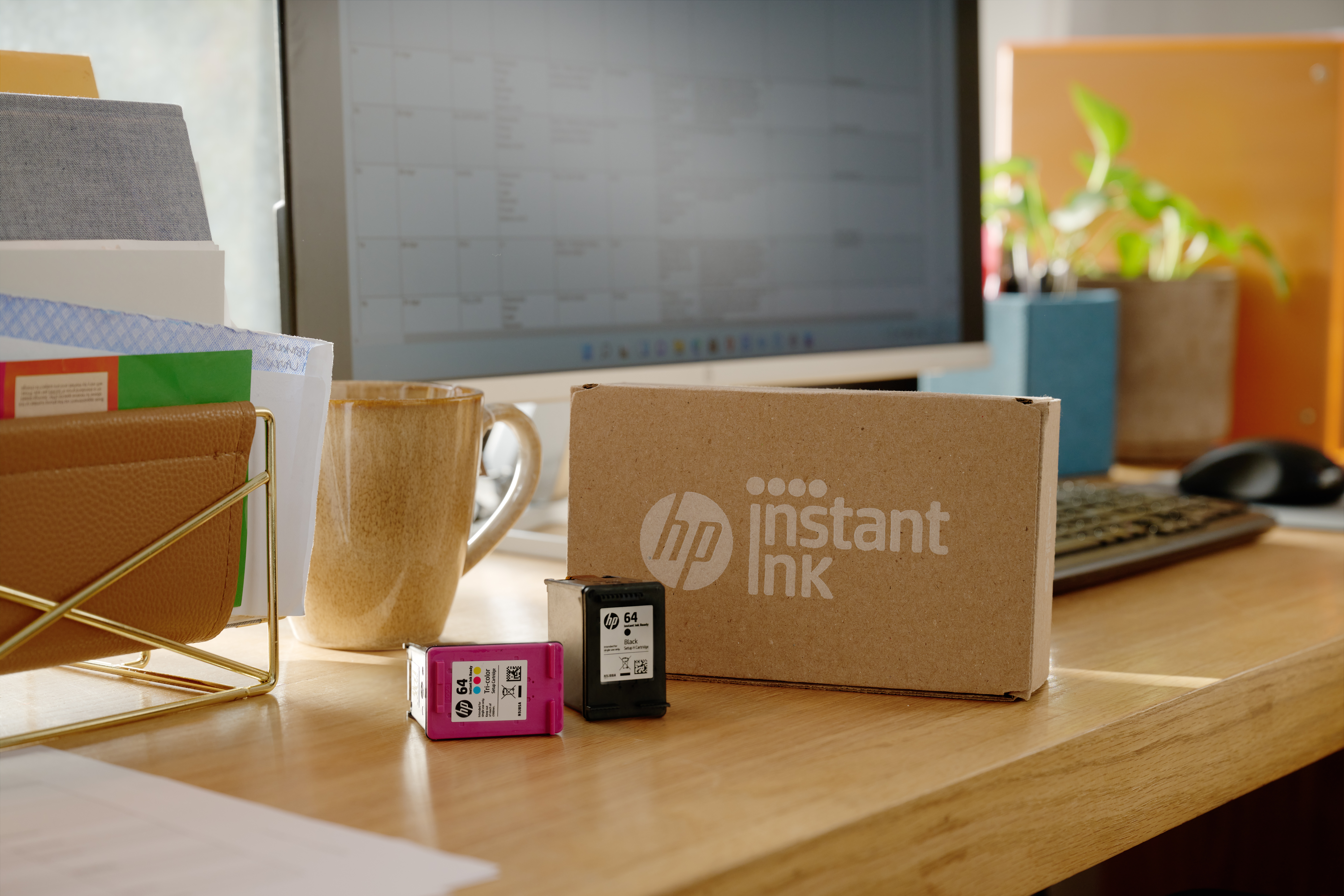 Schreibtisch mit HP Instant Ink-Box, Tintenpatronen, Tasse, Monitor, Dokumenten und einer Pflanze. Die Szene ist gut beleuchtet.