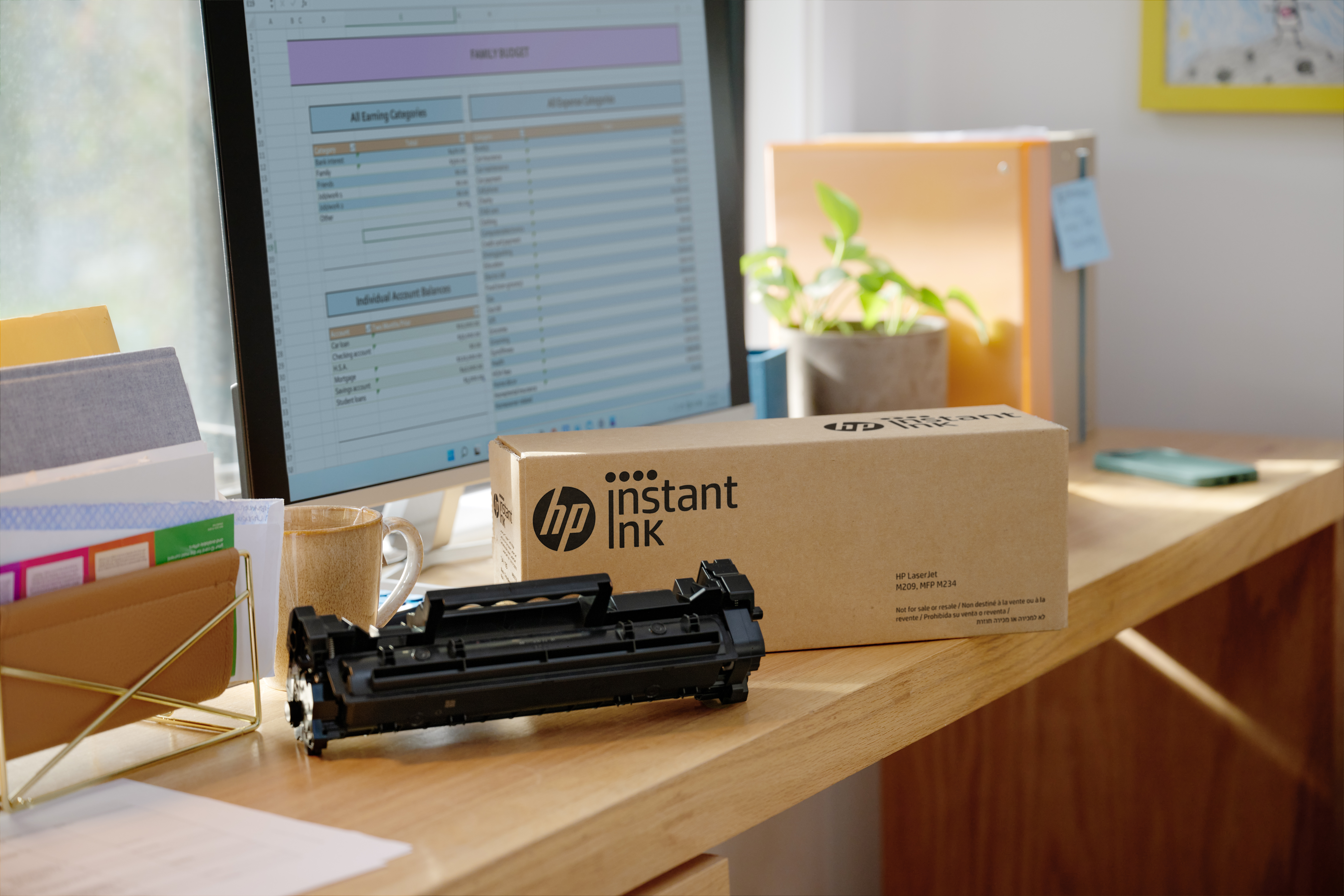 Schreibtisch mit Monitor mit Budget, HP Instant Ink Box, Druckerpatrone und Tasse. Hintergrund: Pflanze.