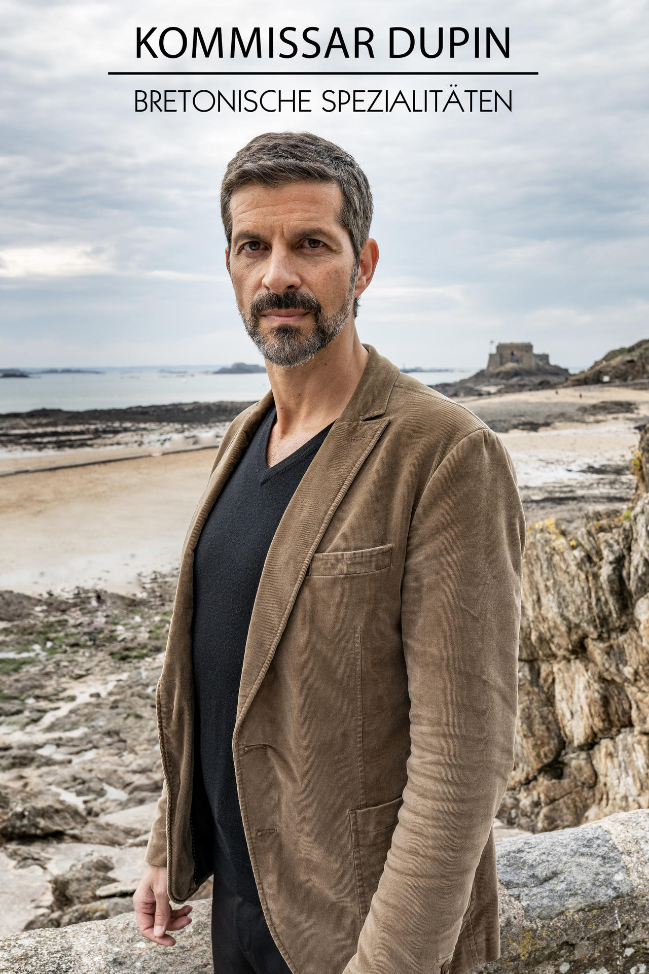 Mann in brauner Jacke steht am Meer. Er hat einen Bart. Im Hintergrund ein Sandstrand und ein Gebäude.