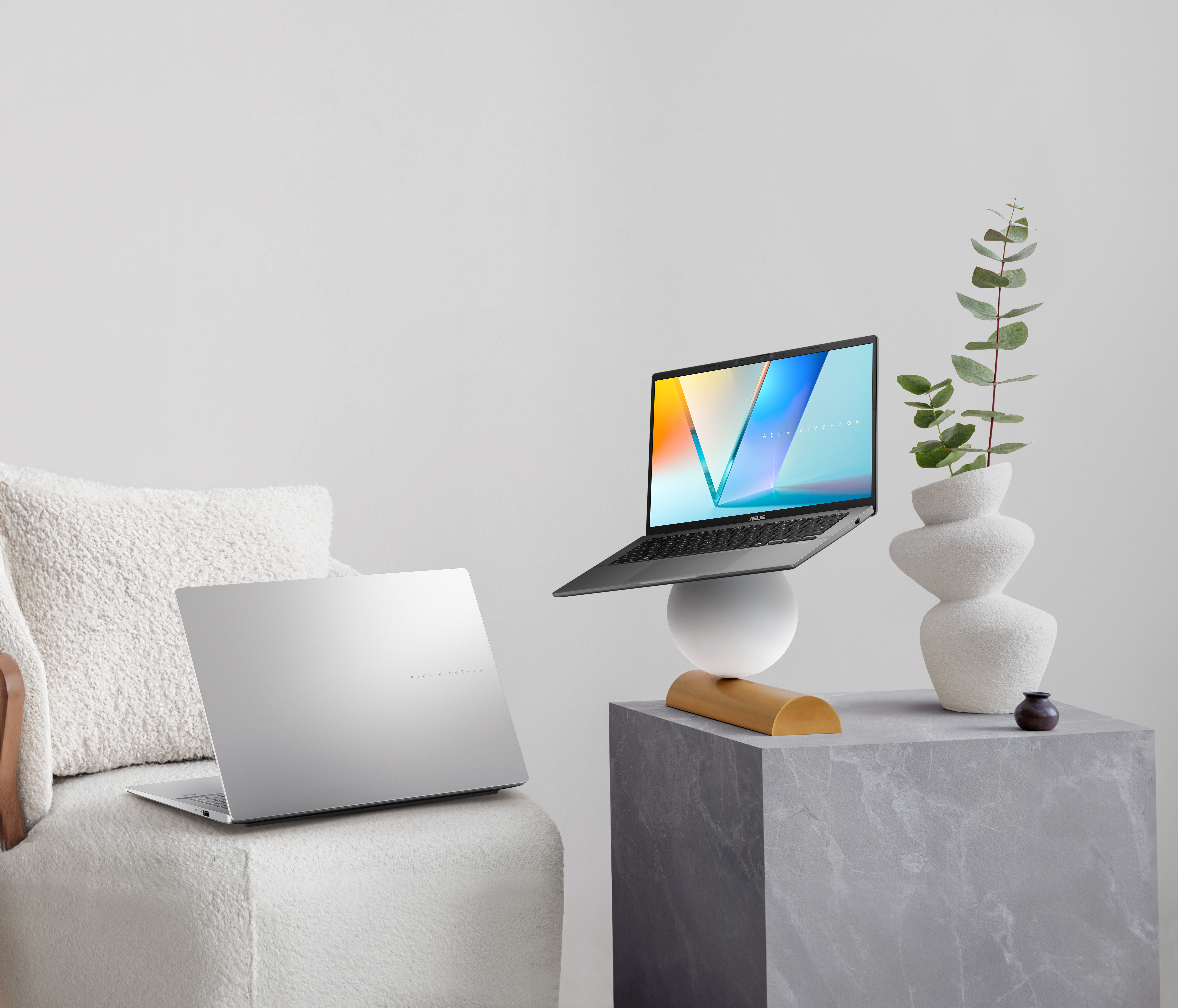 Silberner Laptop auf einer weißen Kugel und Holzsockel, anderer Laptop auf einem weißen Stuhl, Vase mit Pflanze auf einem grauen Würfel.