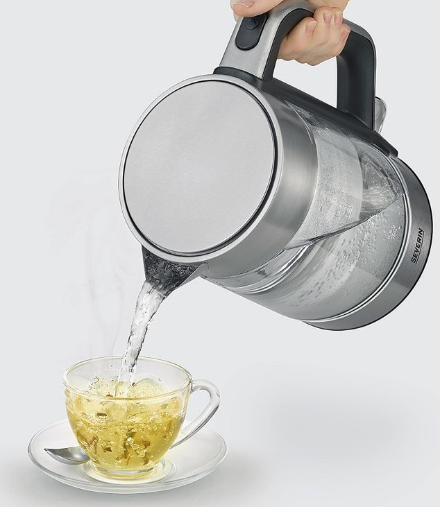 Ręka nalewa wodę z czajnika do filiżanki herbaty. Filiżanka stoi na spodku z łyżeczką.