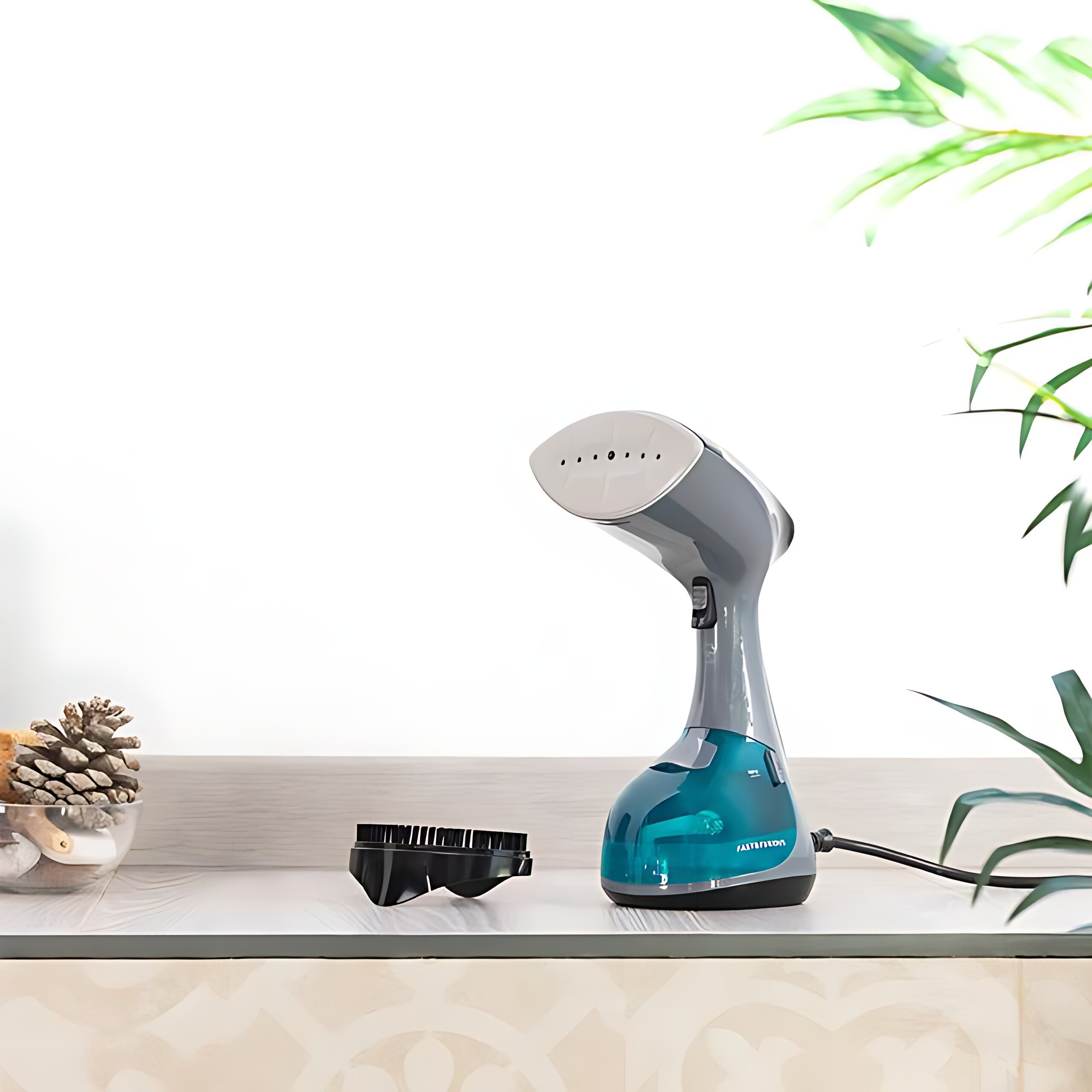 Szaro-turkusowy parownik do ubrań na blacie. Obok szczotka i szyszka w szklanej misce.