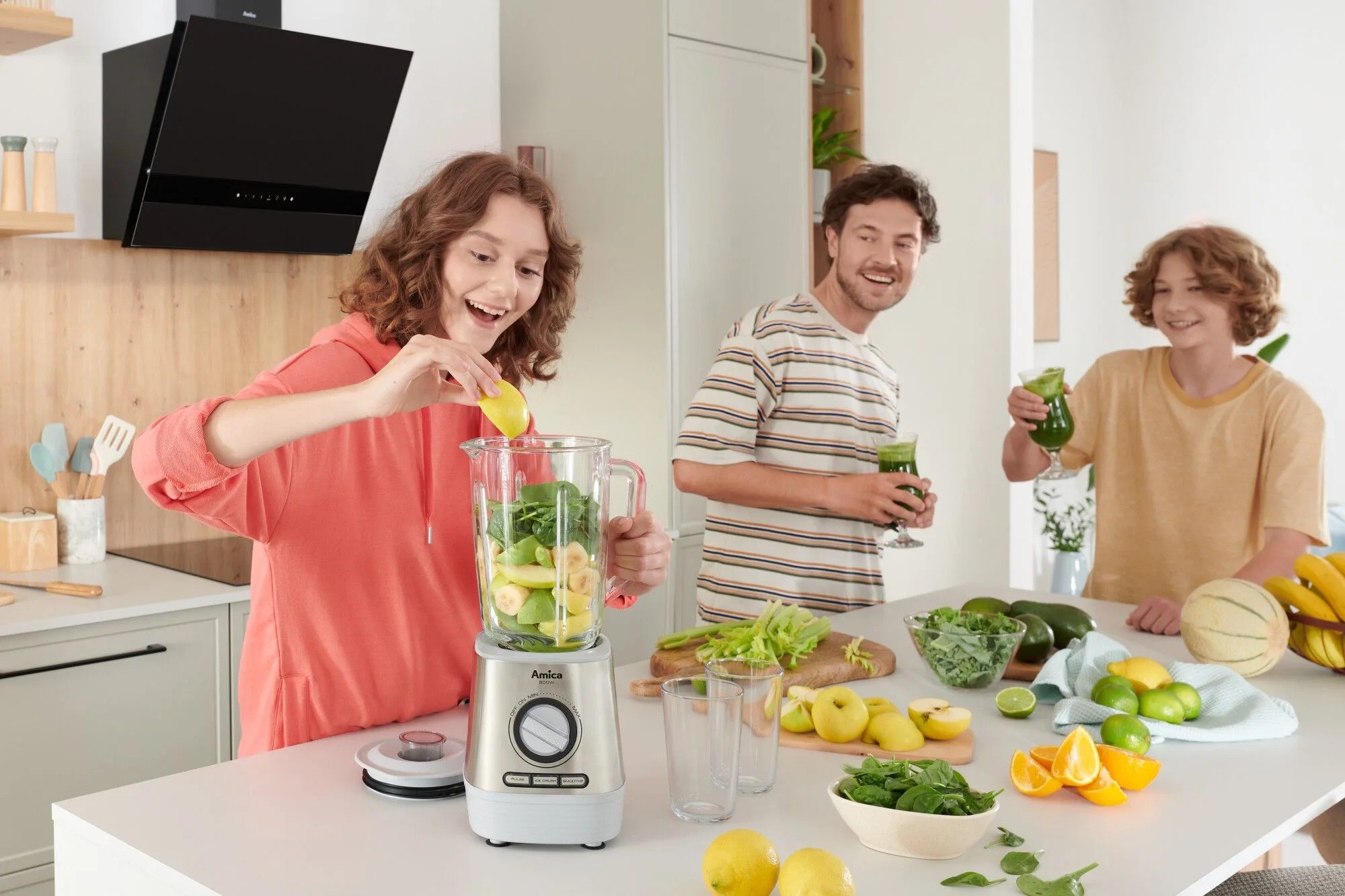 Rodzina robi smoothie z owoców i warzyw. Kobieta dodaje cytrynę do blendera. Dwie osoby trzymają zielone napoje.