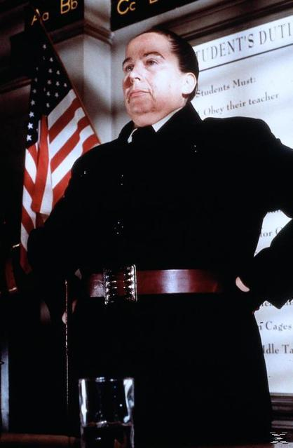 Eine Person in einem schwarzen Mantel steht selbstbewusst da, im Hintergrund eine amerikanische Flagge und ein Schild. Im Vordergrund ein Glas.