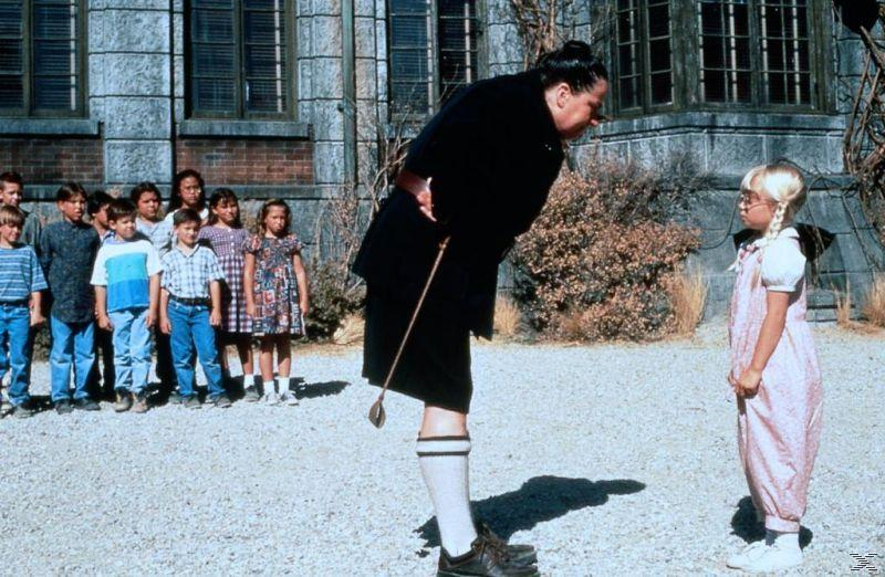 Eine strenge Frau mit Stock steht einem Mädchen gegenüber. Kinder stehen im Hintergrund. Ein Steingebäude bildet die Kulisse.