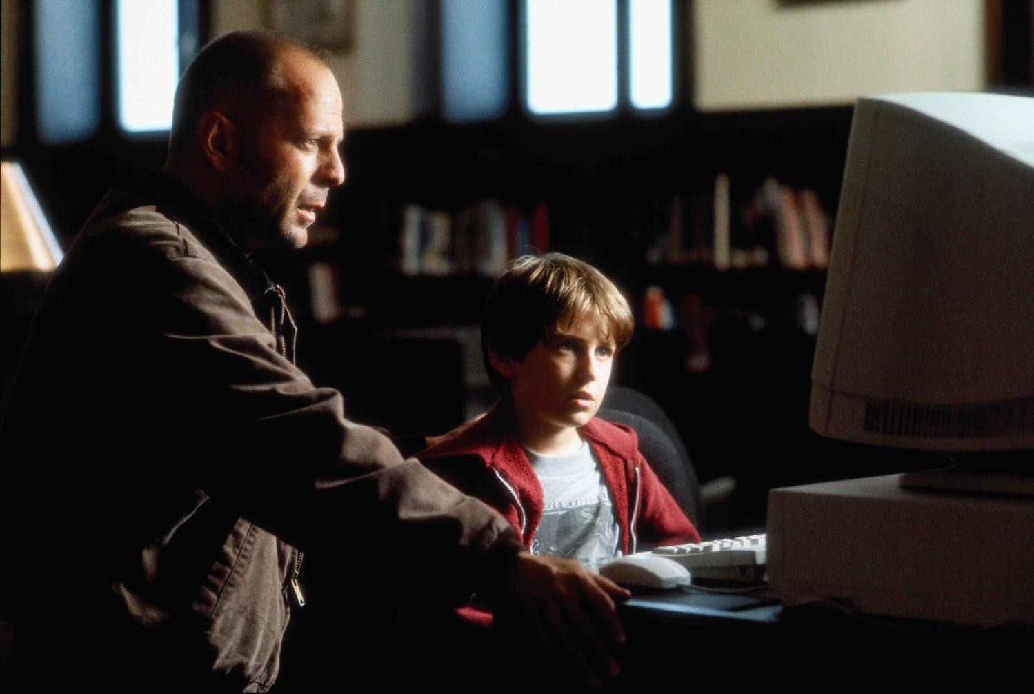 Ein Mann und ein Junge stehen vor einem Computer. Der Junge trägt eine rote Jacke. Im Hintergrund sind Bücherregale.
