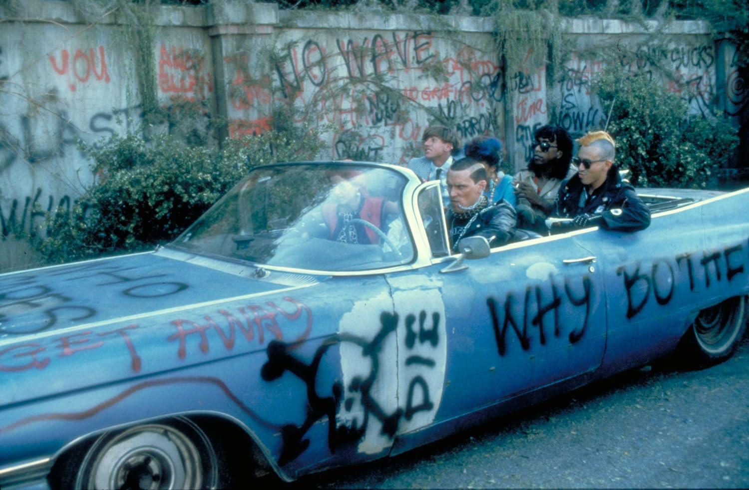 Ein blaues Cabriolet mit Graffiti befördert mehrere Personen. Der Hintergrund ist eine mit Graffiti bedeckte Wand. Die Farbe des Autos ist verblasst.
