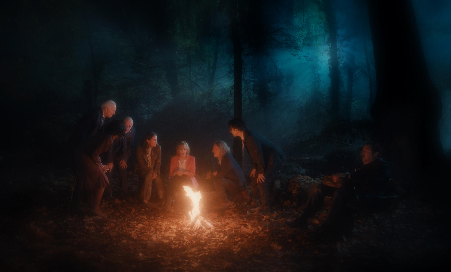 Gruppe von Personen am Lagerfeuer in einem dunklen Wald. Flammen beleuchten Gesichter und Umgebung. Bäume im Hintergrund.