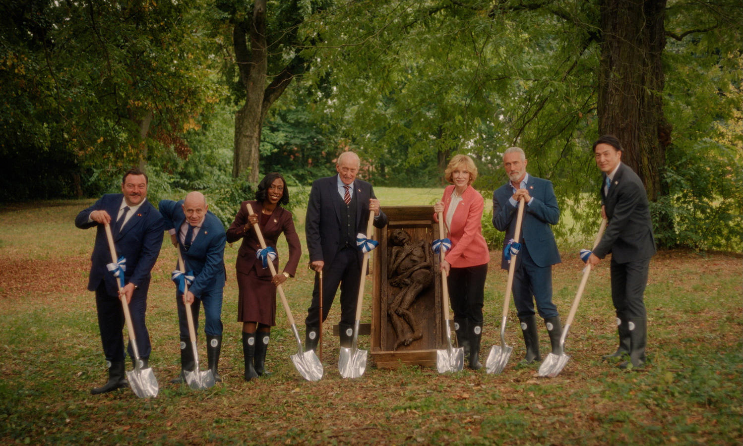 Gruppe von Personen mit Schaufeln, die in der Nähe einer Holzkonstruktion auf einer Wiese graben, Bäume im Hintergrund.