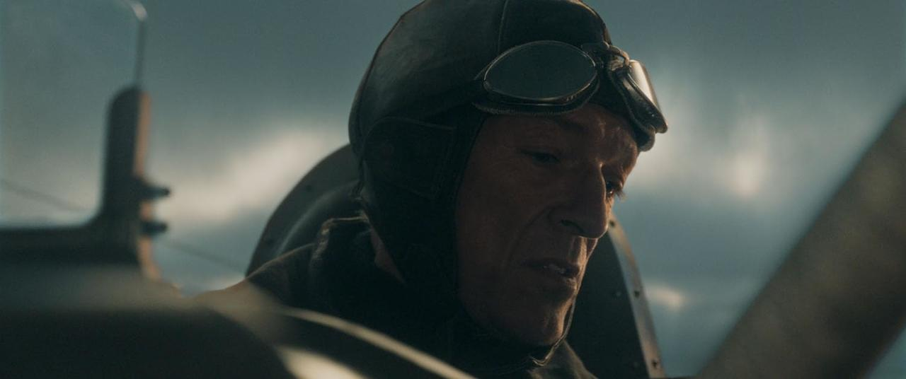 Mann in Fliegerausrüstung, Brille und Helm. Flugzeug sichtbar. Wolkiger Himmel im Hintergrund.