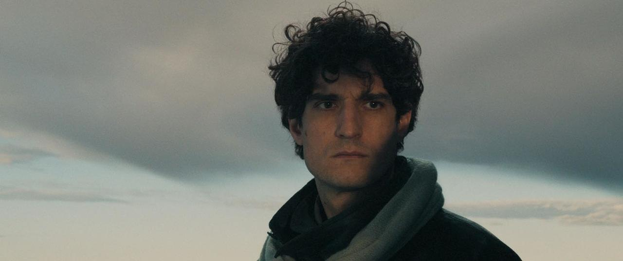 Ein Mann mit lockigem, dunklem Haar und einem Schal blickt den Betrachter an. Der Hintergrund ist ein bewölkter Himmel.