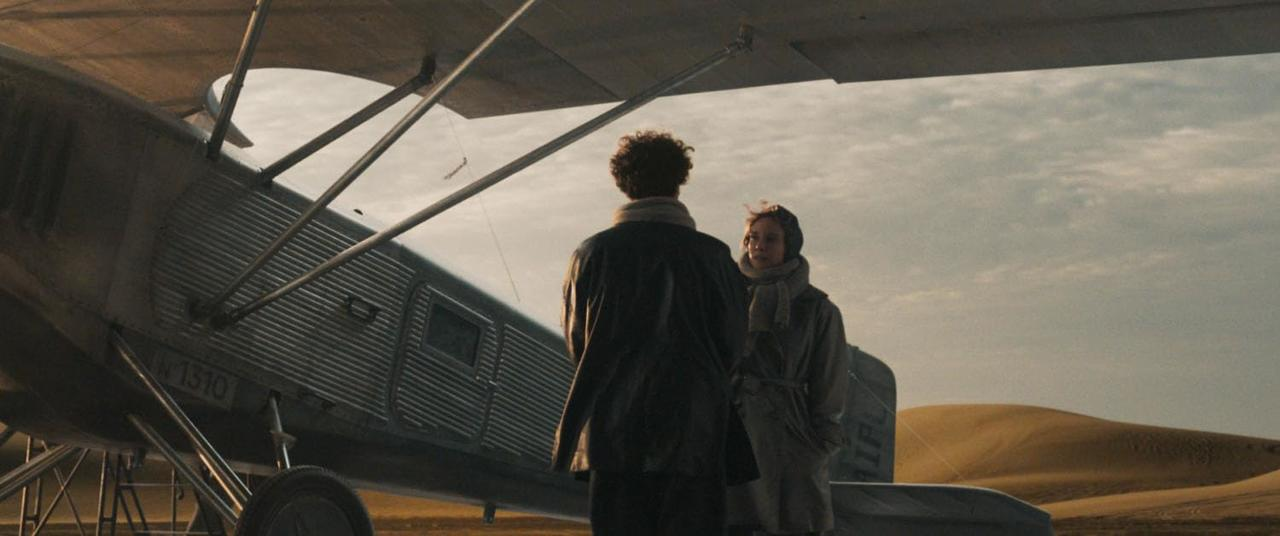 Zwei Personen stehen neben einem alten Flugzeug. Sanddünen befinden sich im Hintergrund unter einem bewölkten Himmel.