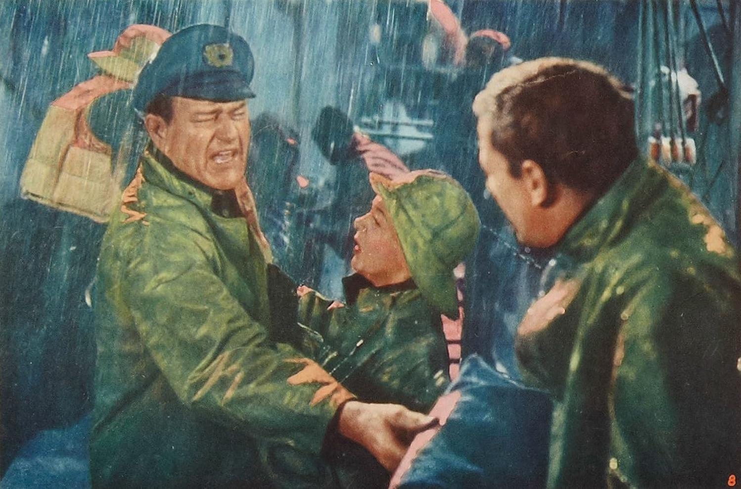 Männer in Regenmänteln kämpfen im Regen. Ein Mann mit Mütze hält ein Kind. Ein anderer Mann schaut zu. Im Hintergrund hängen Rettungswesten.