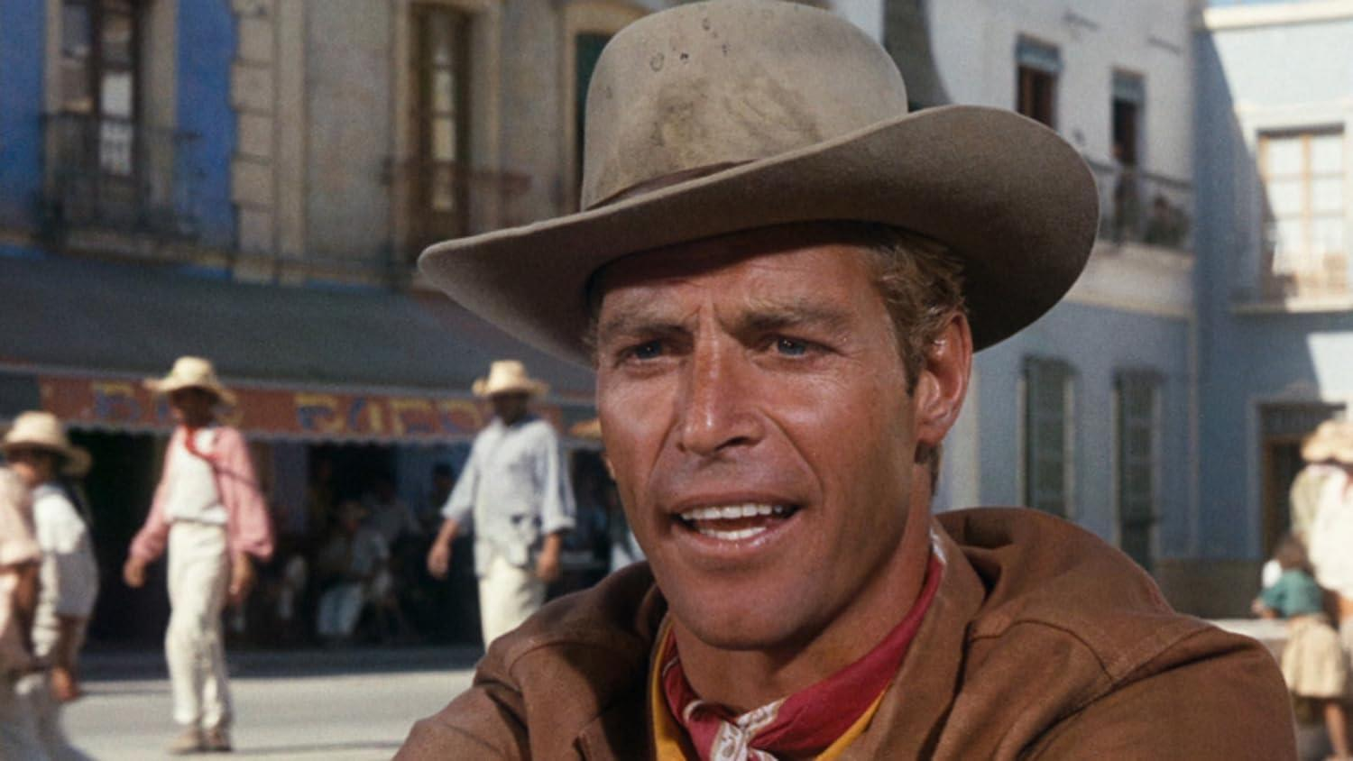 Mann mit Cowboyhut lächelt. Andere mit Hüten und weißer Kleidung im Hintergrund, blaues Gebäude sichtbar.