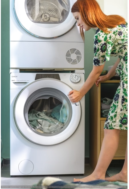 Donna vicino alla lavatrice, asciugatrice impilata sopra. Indossa un vestito floreale. Il bucato è nella lavatrice. Muro verde.