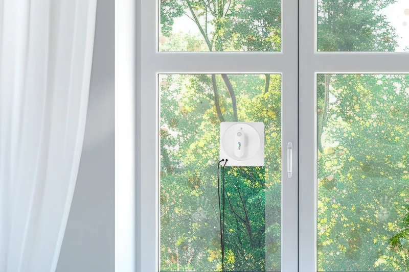 Biały czyścik do okien na szklanym oknie. Zielone drzewa widoczne. Białe zasłony po lewej.