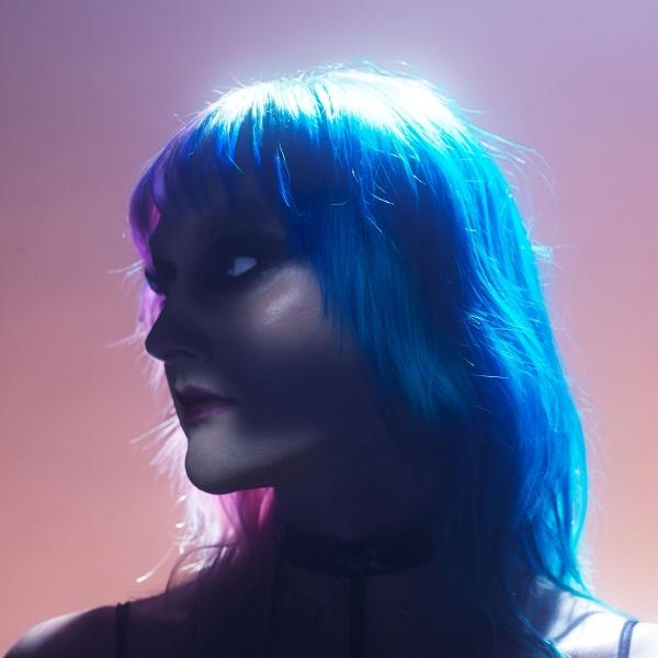 Profil einer Person mit blau-rosa Haaren. Sie trägt einen schwarzen Halsband. Der Hintergrund hat einen rosa-violetten Farbverlauf.