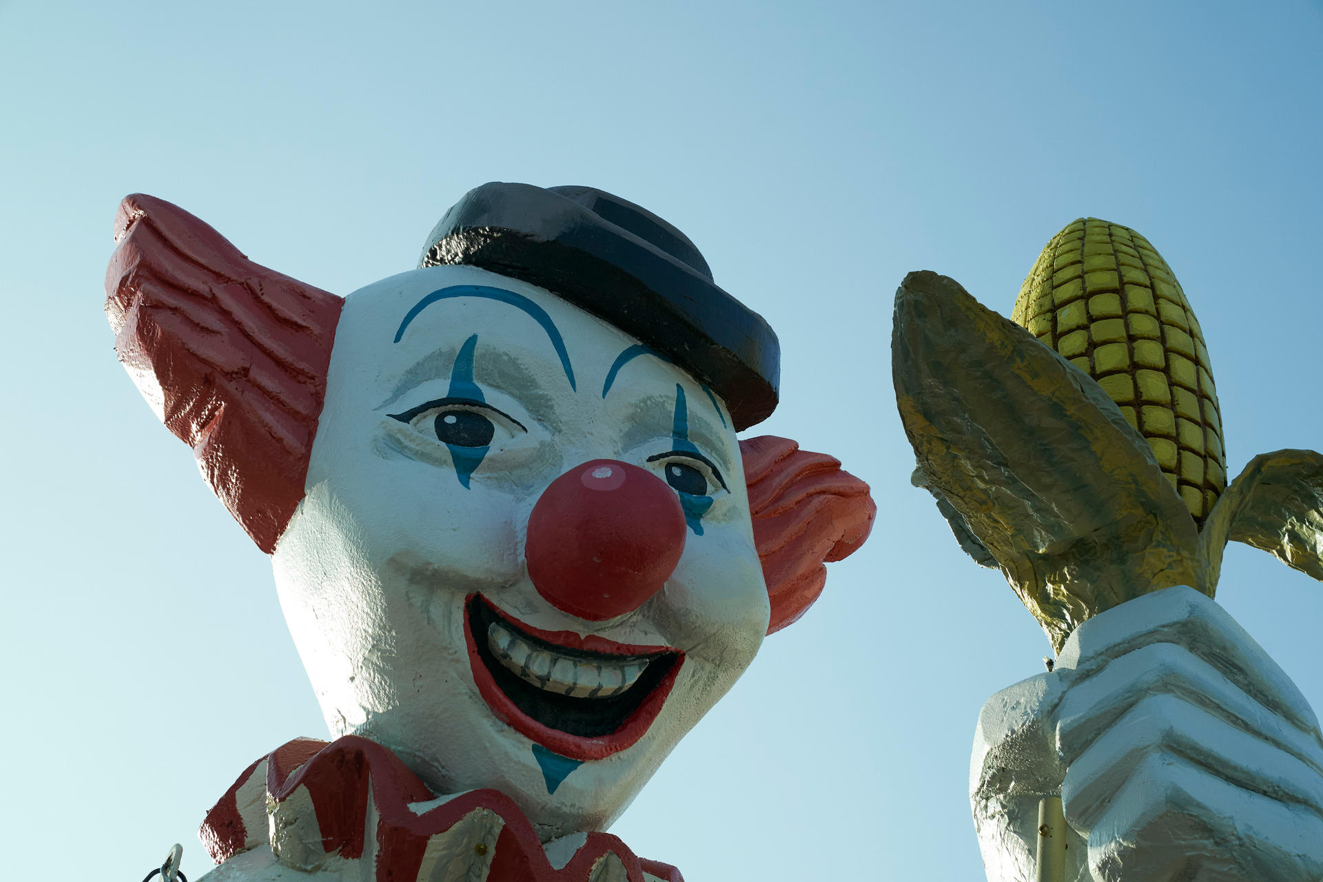 Clownstatue mit roter Nase, Hut und offenem Mund. Sie hält Maiskolben. Der Himmel ist blau.