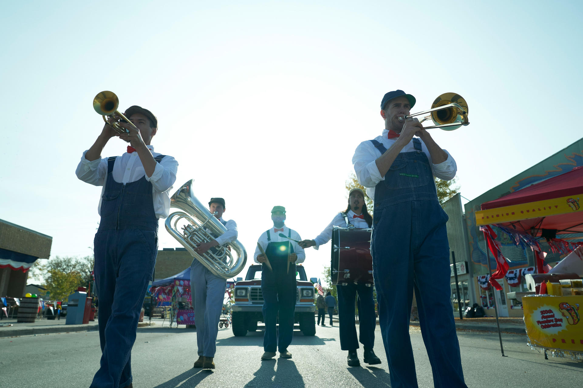Eine Band von Musikern in Overalls spielt Blechblasinstrumente und eine Trommel auf einer sonnigen Straße, mit einem Truck im Hintergrund.
