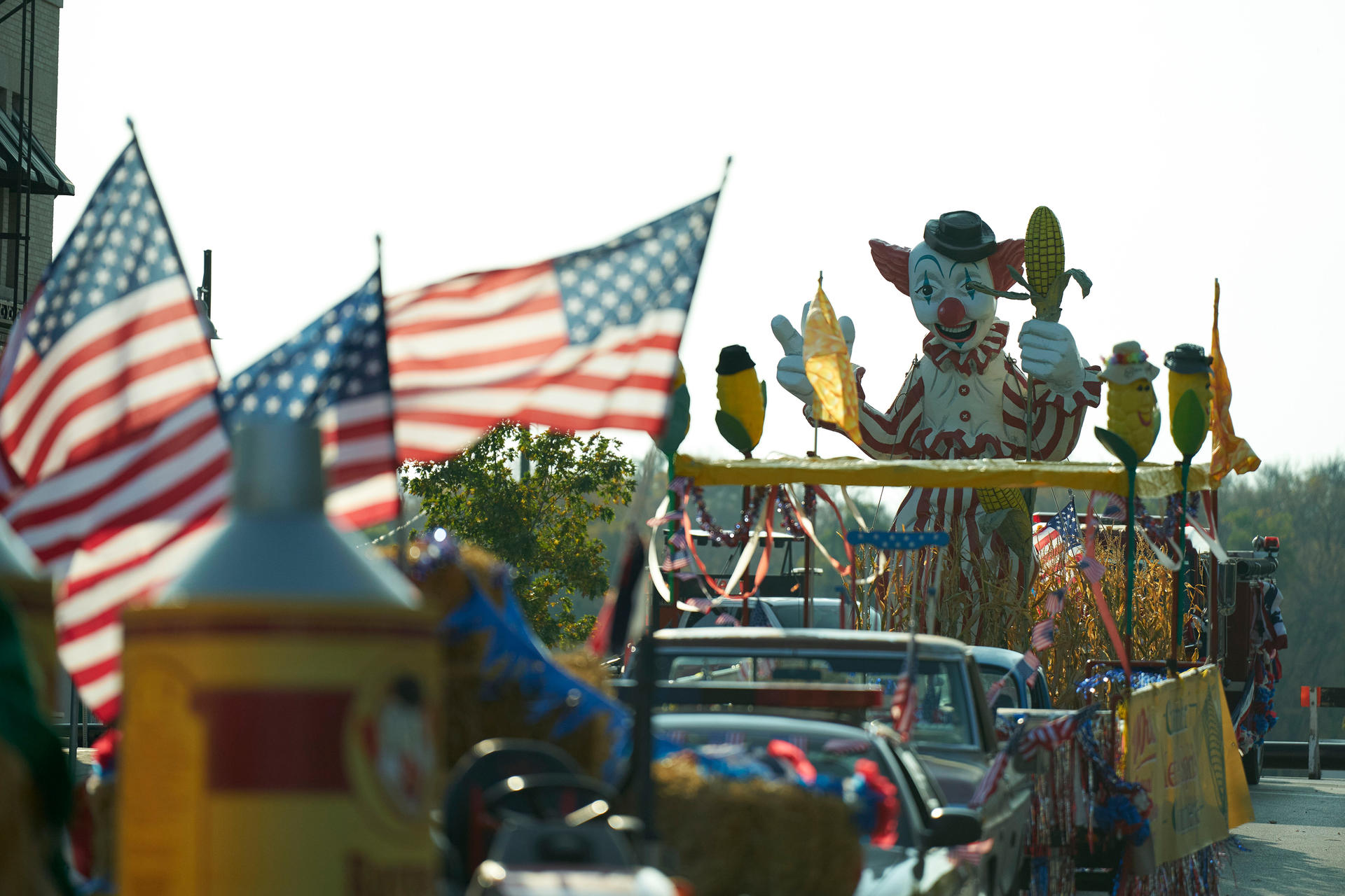 Ein Festwagen mit Clown und amerikanischen Flaggen. Maisdekorationen und Autos füllen die Straße. Heller Himmel im Hintergrund.