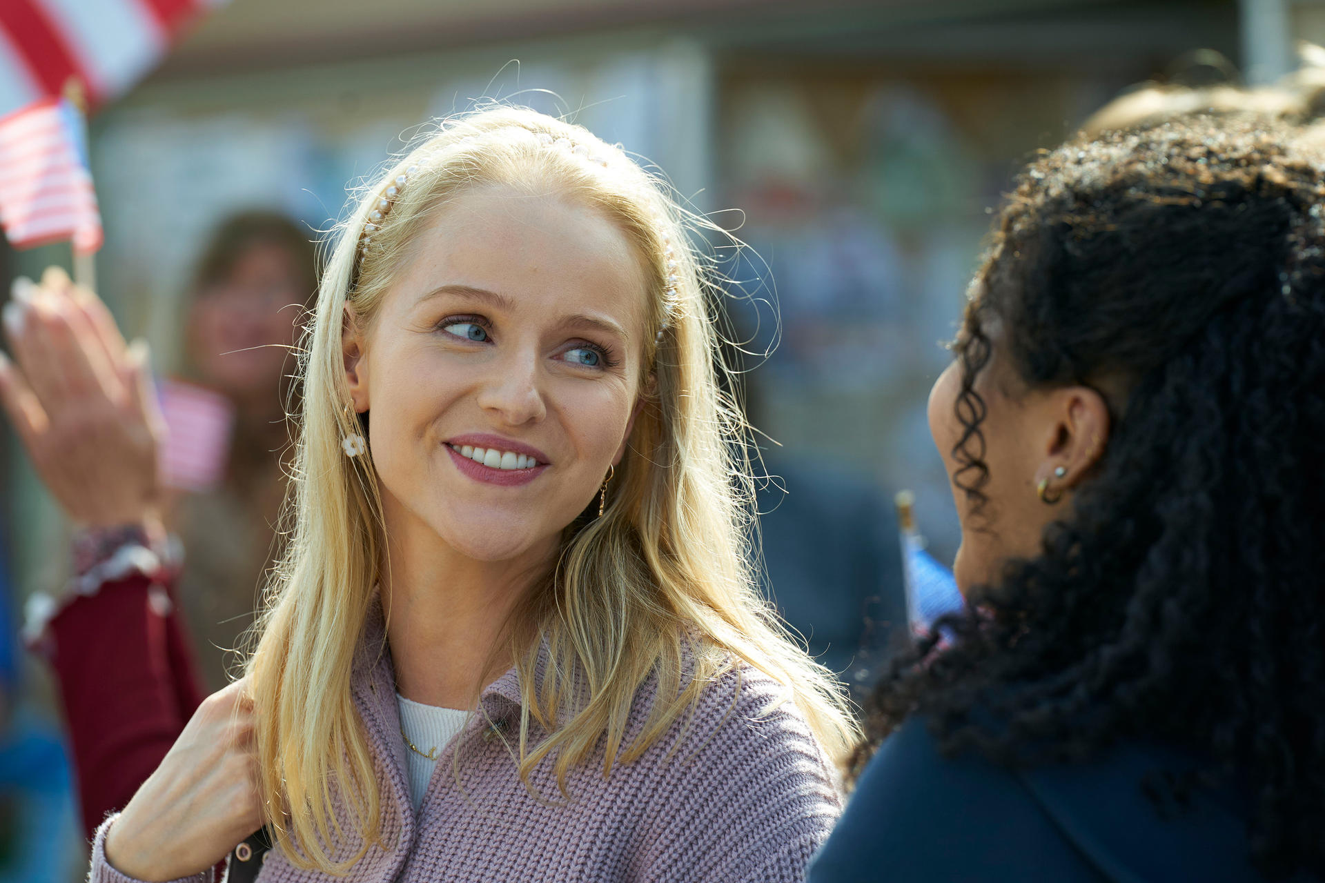 Zwei lächelnde Frauen, eine blonde mit rosa Pullover, die andere mit dunklem, lockigem Haar. Im Hintergrund sind amerikanische Flaggen.
