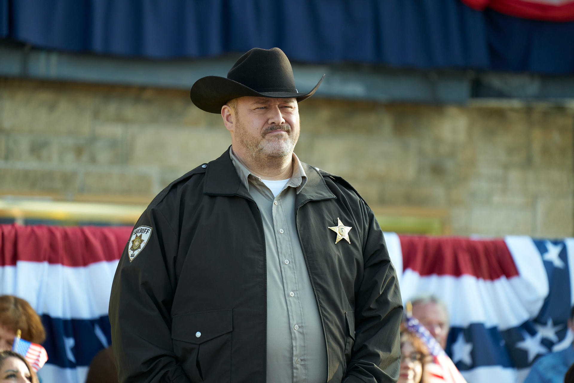 Ein Sheriff mit schwarzem Hut und Jacke. Hinter ihm sind rot-weiß-blaue Dekorationen und eine Steinmauer.