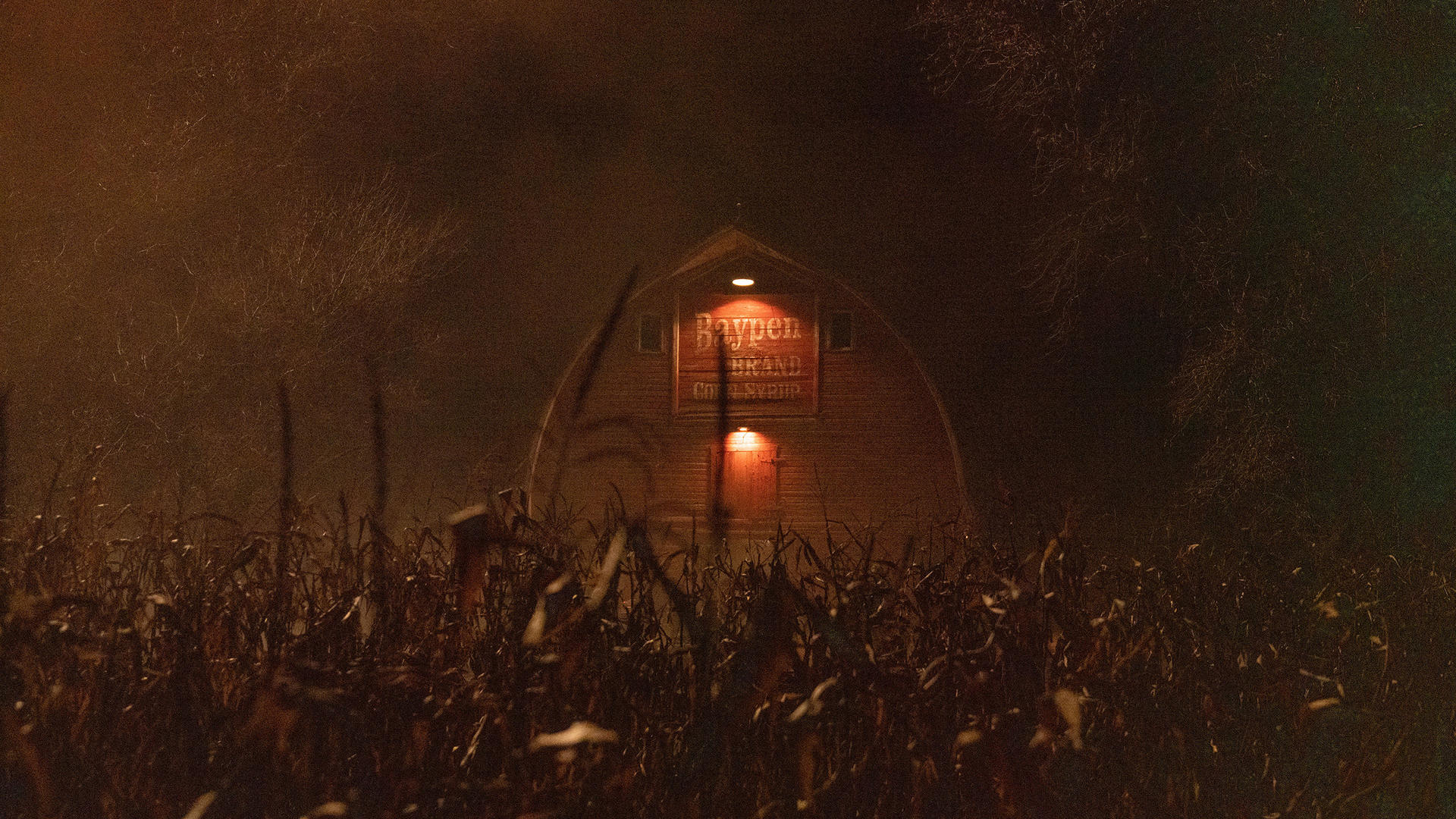 Scheune bei Nacht mit Maisfeld davor. Beleuchtetes Schild mit der Aufschrift "Baypen Brand Corn Syrup". Nebel und gedämpftes Licht erzeugen eine gruselige Atmosphäre.