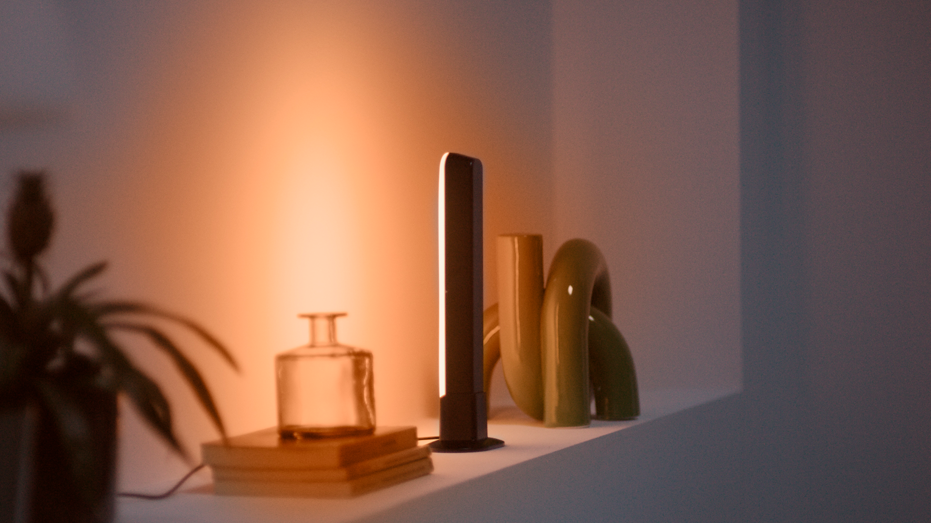 Una lámpara encendida, jarrones, libros y una planta están en un estante blanco, contra una pared cálida e iluminada.