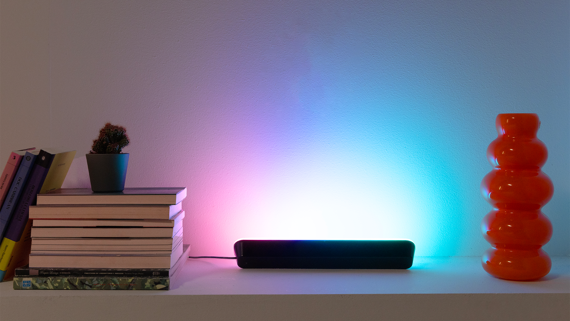 Hay libros, un cactus y un jarrón naranja en una estantería. Una barra de luz negra emite luz de colores contra una pared blanca.