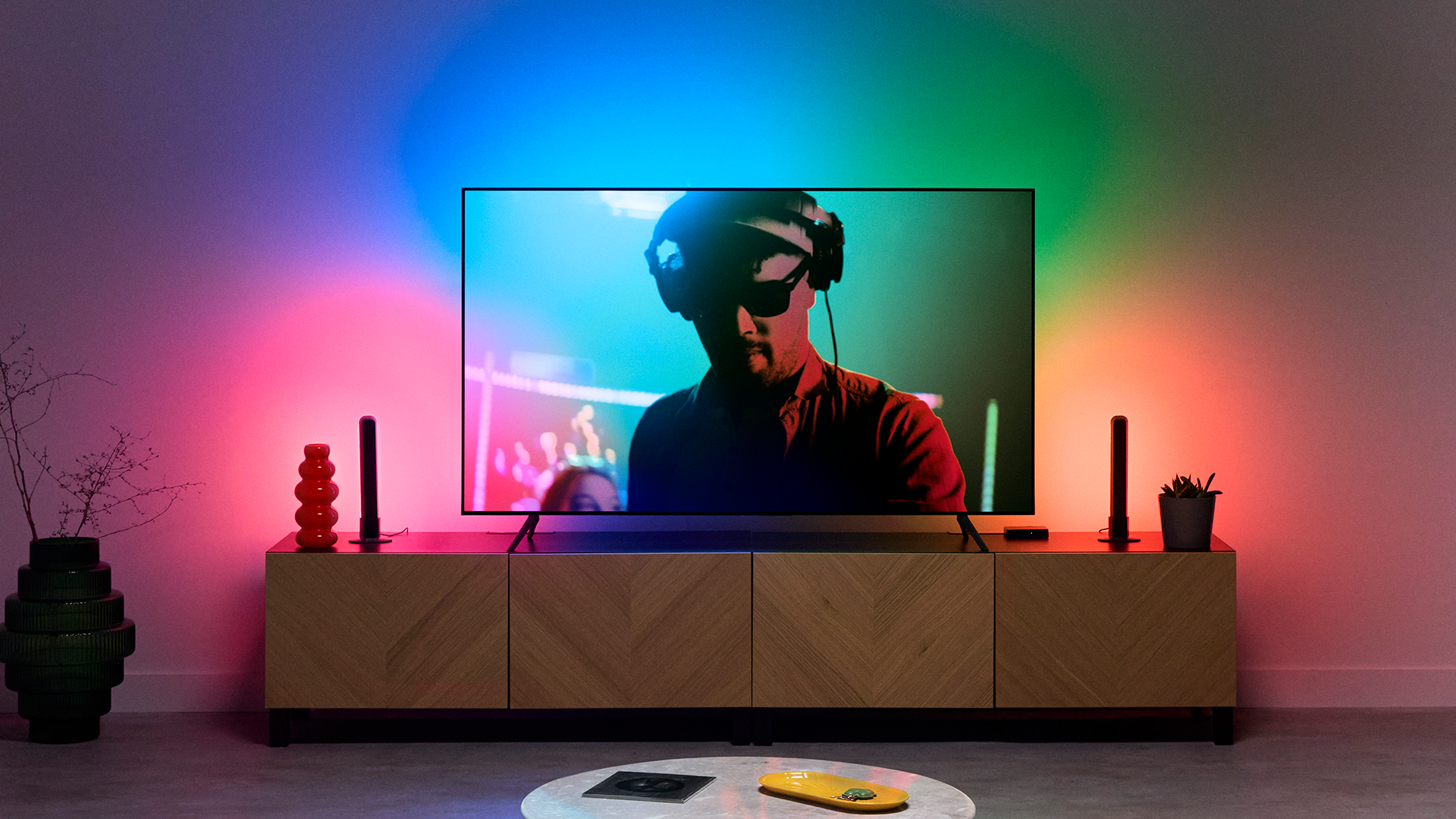 Un hombre con auriculares está en la pantalla de un televisor. La habitación tiene iluminación colorida y un mueble de madera con objetos decorativos.
