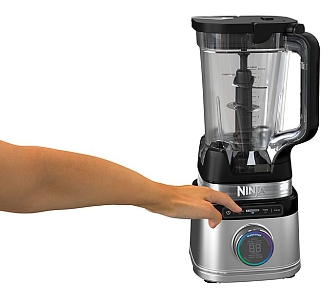 Ręka naciska przycisk na blenderze Ninja. Blender jest srebrny i czarny, z przezroczystym pojemnikiem. Tło jest białe.