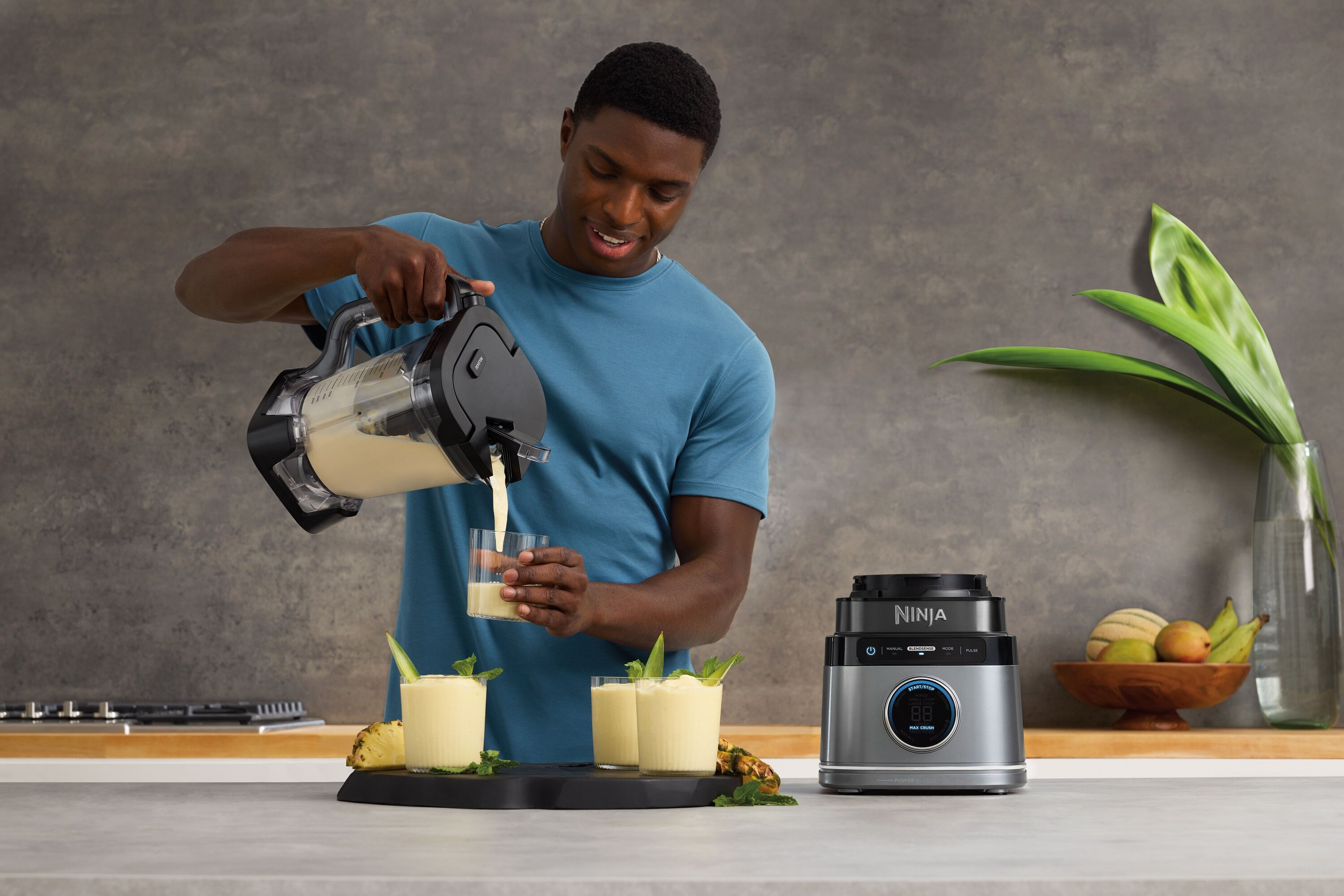 Mężczyzna nalewa smoothie z blendera do szklanki. Ananas i napoje na tacy, blender po prawej.