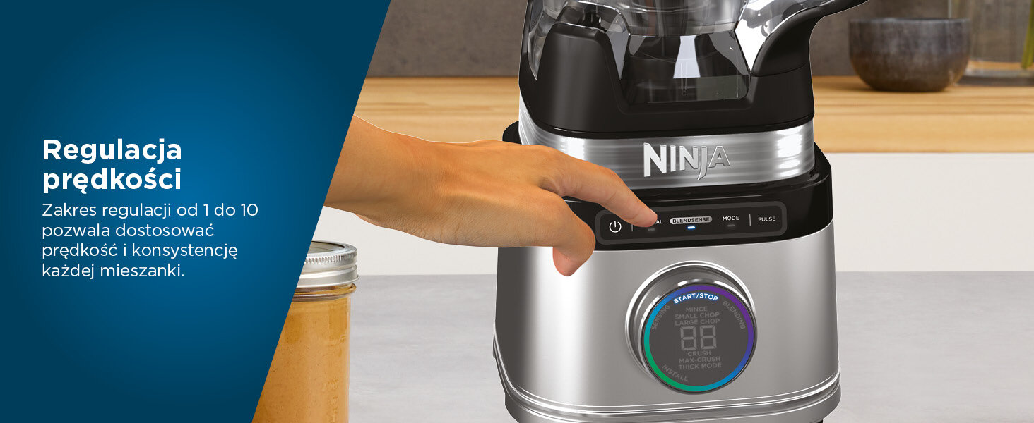 Ręka naciska przycisk na blenderze Ninja. Blender stoi na jasnej powierzchni. Słoik z zawartością znajduje się na pierwszym planie.