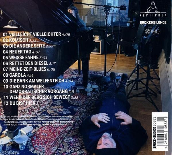 Mann liegt auf dem Boden und blickt nach oben. Klavier, Mikrofon und Musik-Trackliste im Hintergrund. CD-Cover mit Logo und Name.