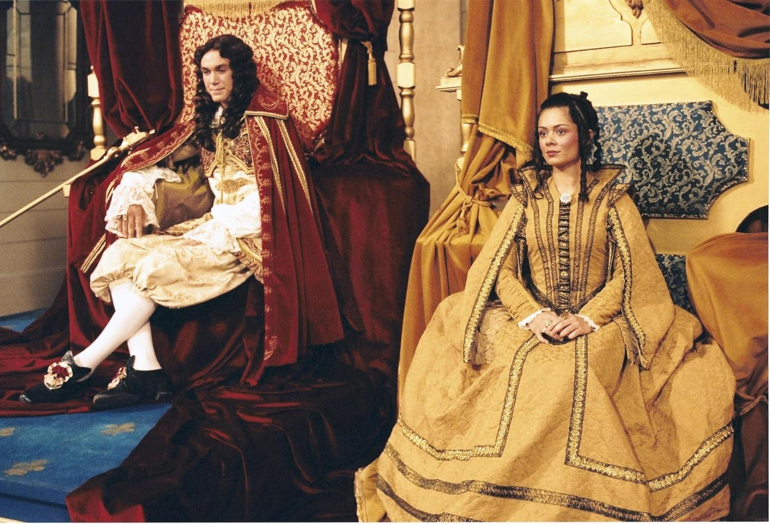 Ein Mann und eine Frau in historischen Kostümen sitzen auf Thronen. Der Mann trägt einen roten Umhang. Die Frau trägt ein goldenes Kleid.