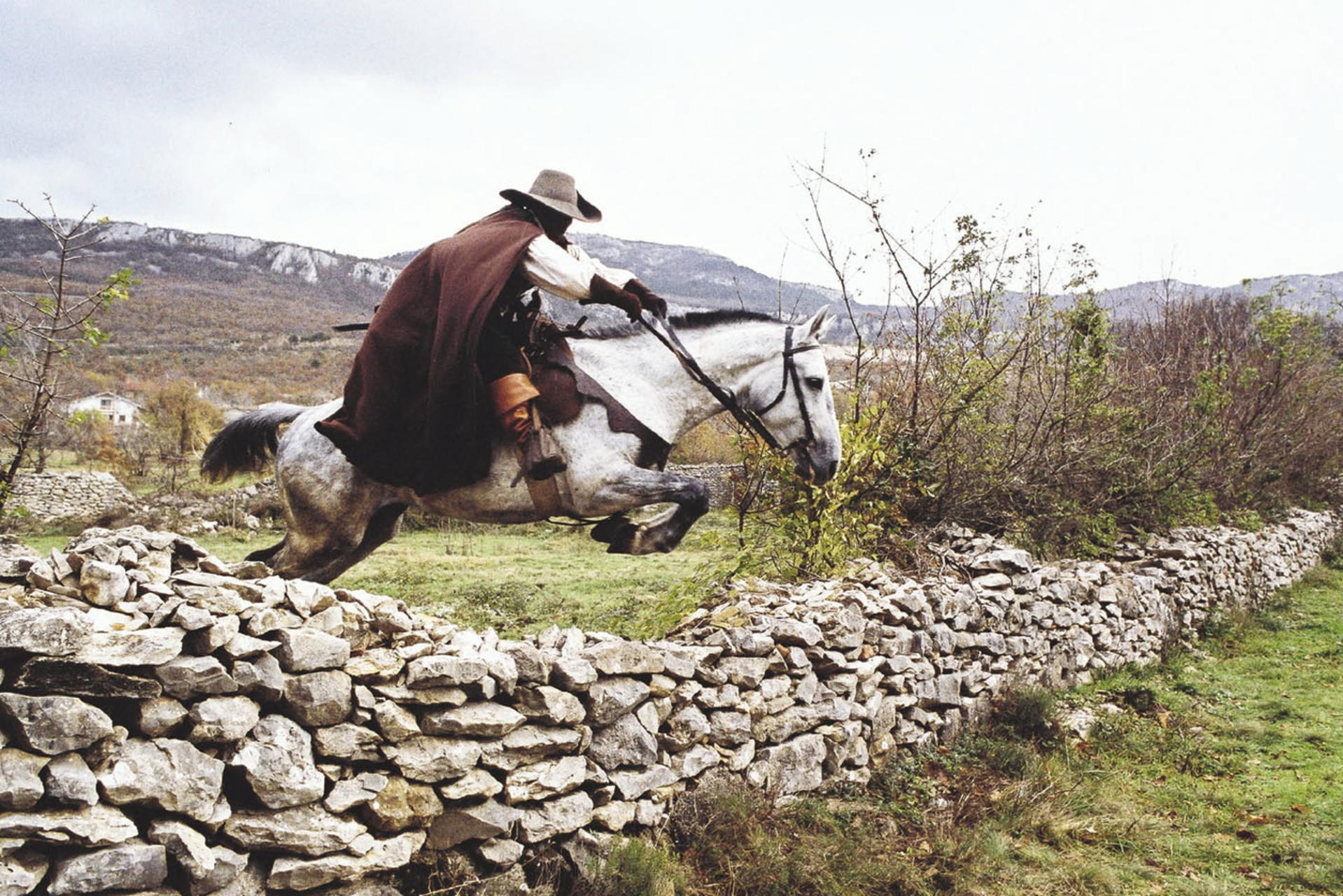 Mann auf Pferd springt über Steinmauer. Pferd ist weiß, Mann trägt Umhang. Hintergrund Berge, Gras und bewölkter Himmel.