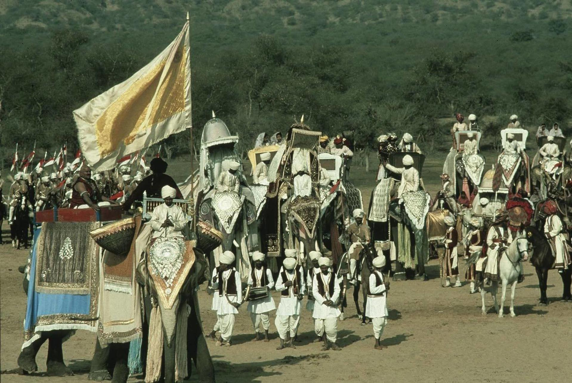 Eine indische Parade mit Elefanten und Pferden. Menschen in traditioneller Kleidung reiten auf den Tieren, mit einer großen Flagge.
