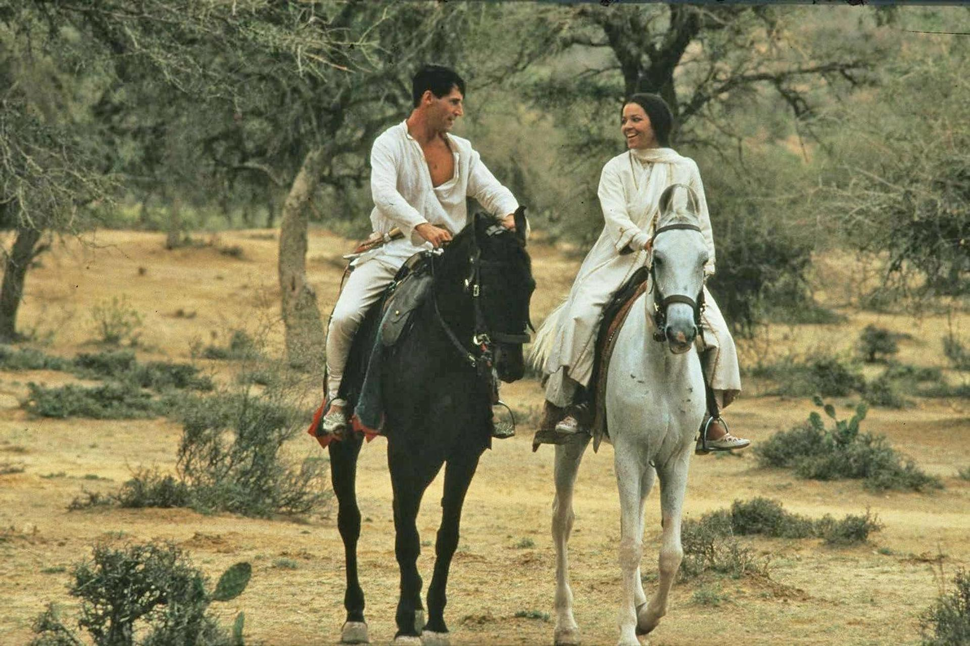 Zwei Personen auf Pferden, einem schwarzen und einem weißen, reiten durch ein braunes Feld. Bäume im Hintergrund.