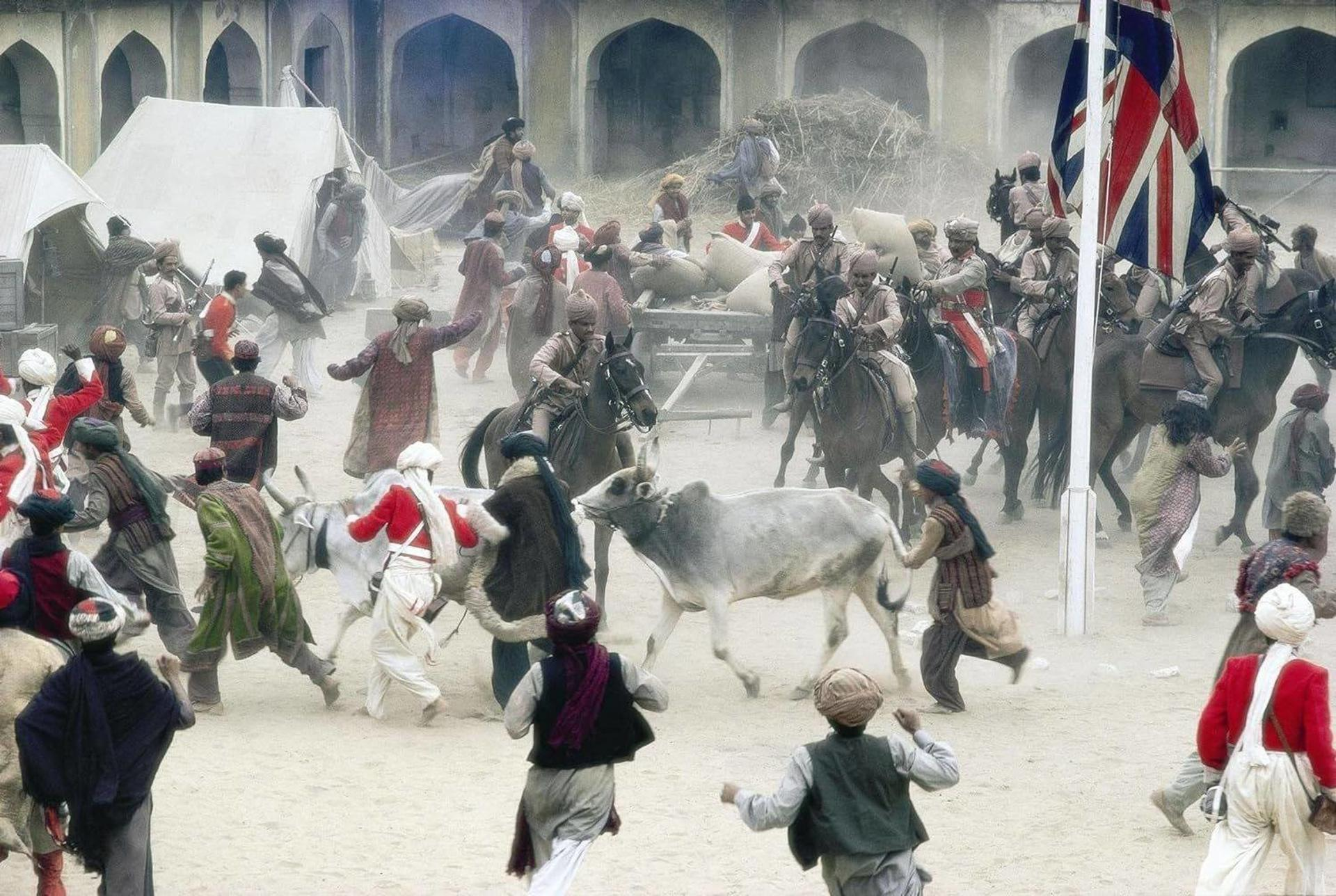 Szene zeigt ein historisches Ereignis mit rennenden Menschen, Pferden, Zelten und einer britischen Flagge. Einige Figuren tragen rote Mäntel.