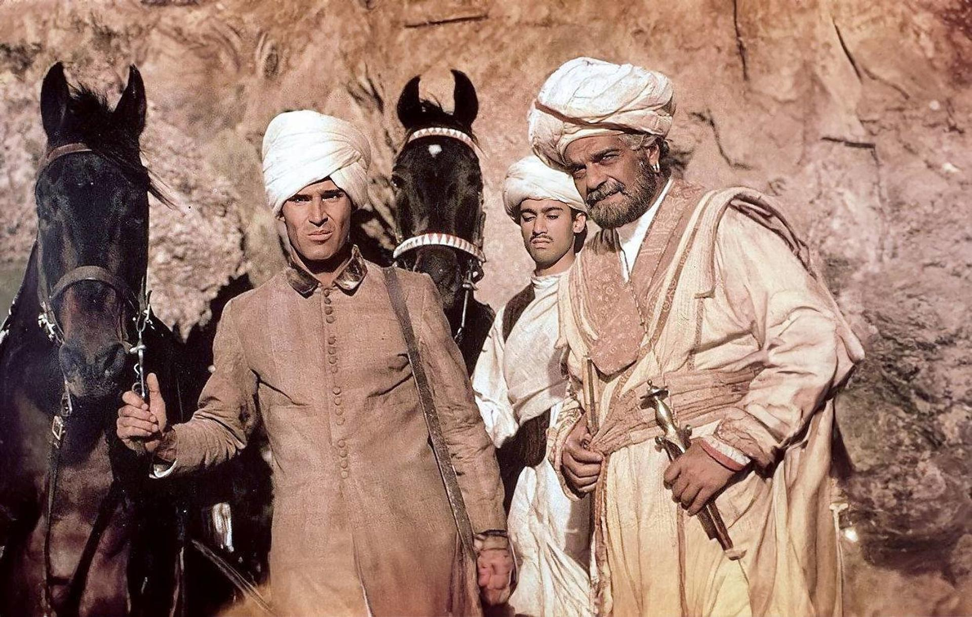 Männer in Turbans und Roben stehen mit Pferden. Einer hält ein Schwert. Der Hintergrund ist eine Steinmauer.