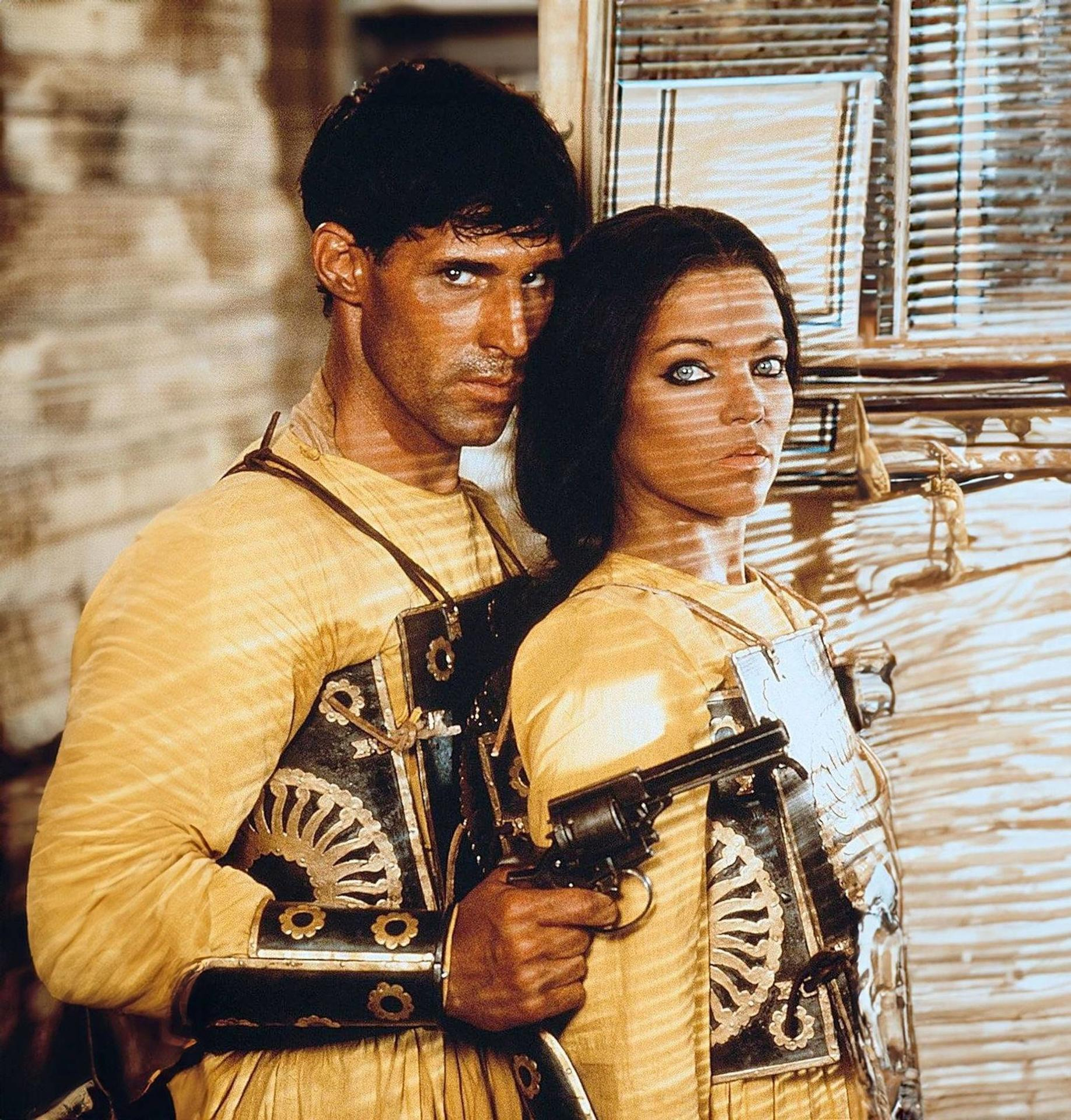 Mann und Frau in gelben Outfits, Mann hält eine Pistole. Beide stehen vor einer Holzwand.