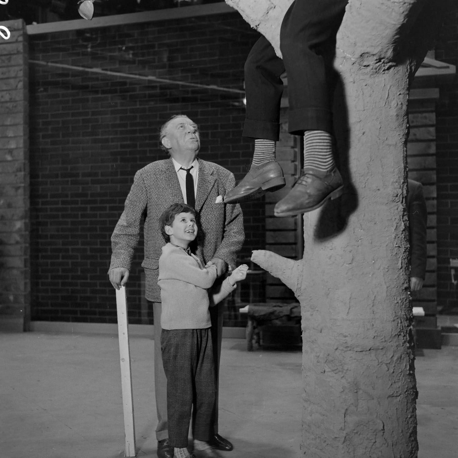 Mann und Kind blicken auf die Beine einer Person in einem Baum. Im Hintergrund eine Backsteinmauer und eine weitere Person.