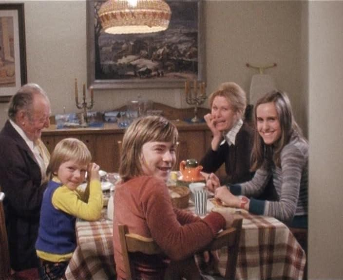 Familie am Tisch. Ein Großvater, zwei Kinder und zwei Frauen sitzen. Im Hintergrund ein Gemälde und eine Lampe.