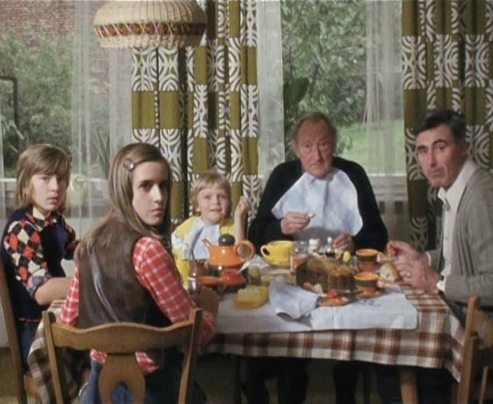Familie am Tisch. Leute essen. Tisch hat Kuchen und Teeservice. Karierte Tischdecke. Gardinen im Hintergrund.
