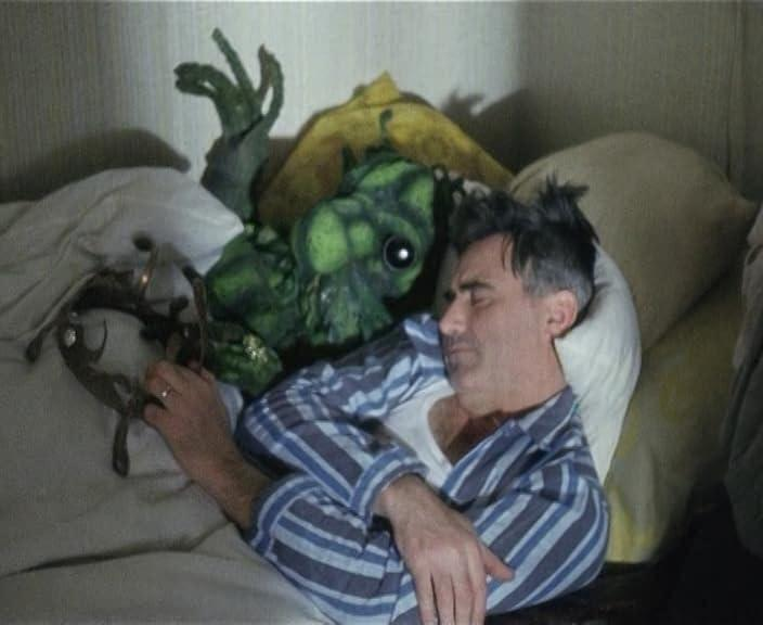 Mann im gestreiften Schlafanzug schläft im Bett. Eine grüne Alien-Puppe ist neben ihm. Bettwäsche und Kissen umgeben sie.