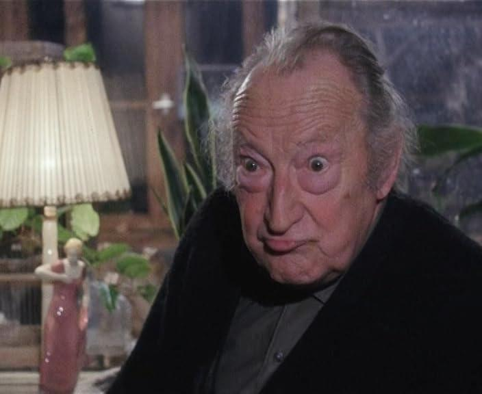 Ein älterer Mann mit aufgerissenen Augen und überraschtem Gesichtsausdruck. Er trägt ein schwarzes Hemd. Im Hintergrund sind eine Lampe und Pflanzen.